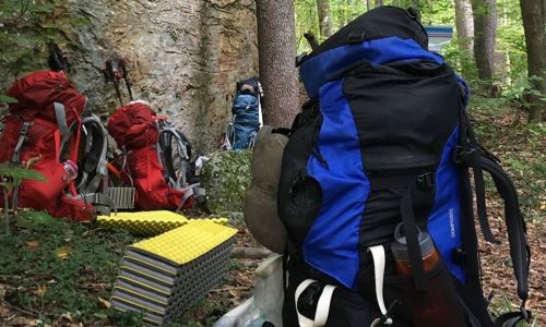 A close-up of a large backpack in a forest, with other backpacks and sleeping pads visible in the background.