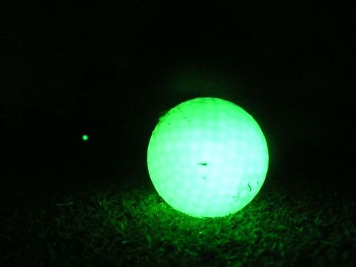 A glowing green golf ball sits on grass in a dark setting, with a faint light from another ball in the background.