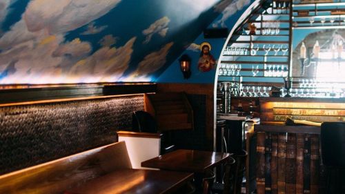 Cozy interior of a bar with cloud-painted ceiling, wooden tables, and shelves lined with glasses.