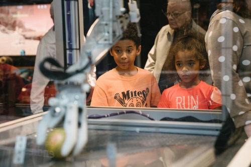 Two children watch a robotic arm interact with an object, with adults in the background at a science exhibit.