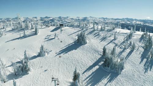 A snowy mountain landscape with ski lifts, trees, and skiers under a clear blue sky. Shadows stretch across the snow.