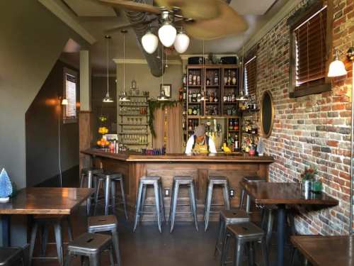 A cozy bar with wooden furniture, a well-stocked shelf, and a bartender preparing drinks in a warm, inviting atmosphere.