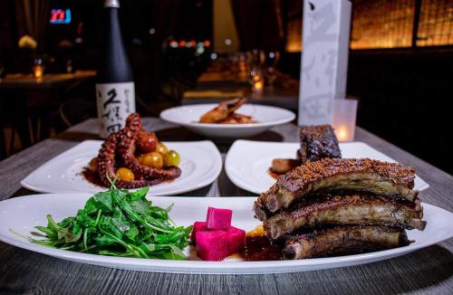 A plate of succulent ribs with greens and pink pickled cubes, with octopus and other dishes in the background.