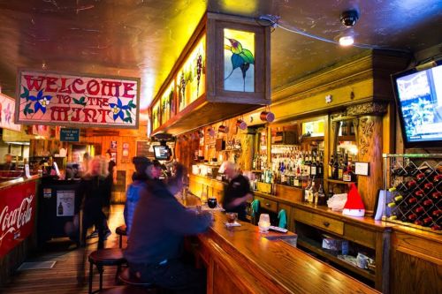 A lively bar scene with colorful decor, a wooden counter, and patrons enjoying drinks in a warm, inviting atmosphere.