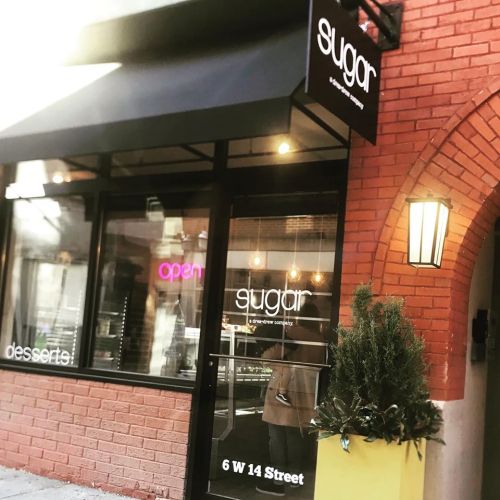 Exterior of a dessert shop named "sugar," featuring a black awning and a bright sign, with an open door.