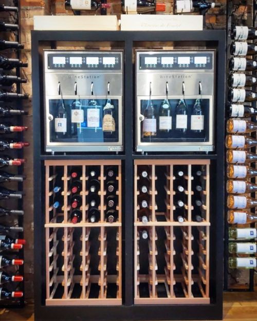 A wine station with two dispensers and a wooden rack, surrounded by bottles of wine on the wall.