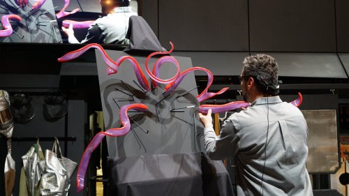 A person interacts with a large, abstract sculpture featuring red, twisting forms against a dark backdrop.