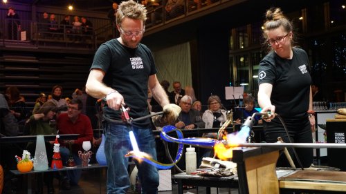 Two glassblowers demonstrate their craft, using torches to shape molten glass in a lively workshop setting.