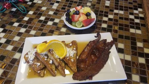 A plate of French toast with syrup and orange slices, alongside crispy bacon and a bowl of mixed fruit.
