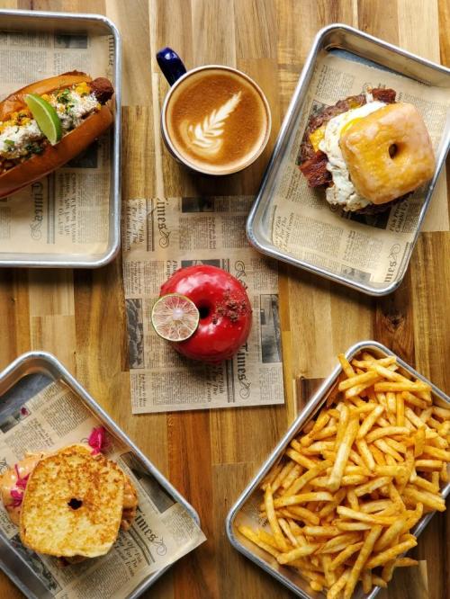 A flat lay of a hot dog, a sandwich with a donut, fries, a red donut, and a latte on newspaper-lined trays.