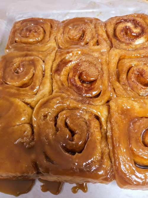 A close-up of freshly baked cinnamon rolls drizzled with caramel glaze, arranged in a square on parchment paper.