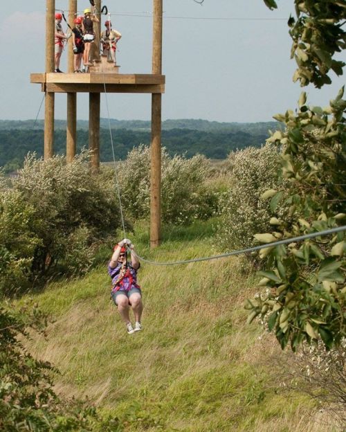 A person zip-lining through a grassy area, with a platform and others visible in the background.