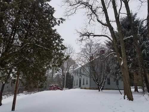 A snowy landscape featuring a gray house surrounded by trees, with a wooden cart visible in the background.