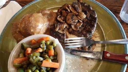 A plate with a grilled steak topped with mushrooms, mashed potatoes with gravy, and a side of mixed vegetables.