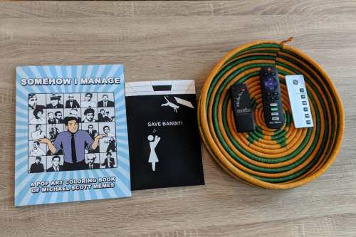 A coloring book of Michael Scott memes, a black and white flyer, and remote controls on a woven basket.