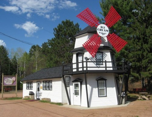 A charming ice cream shop with a windmill on top, surrounded by trees and a gravel path.