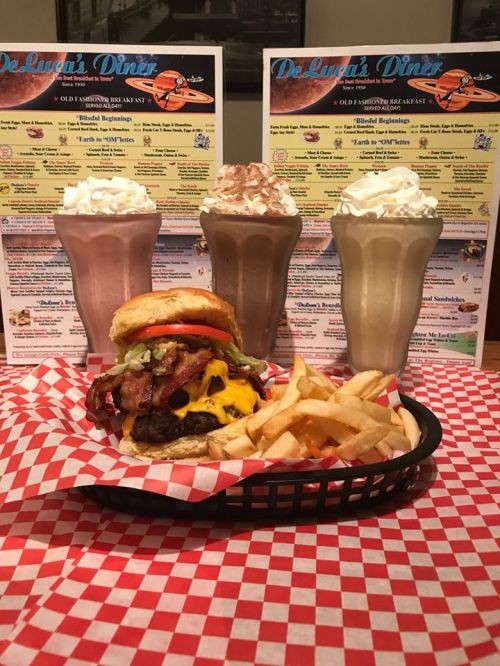 A juicy burger with bacon and cheese, served with fries and three milkshakes in the background at De Luca's Diner.