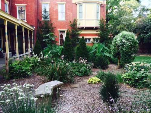 Lush garden with various plants, trees, and a stone bench, set against a red brick building.