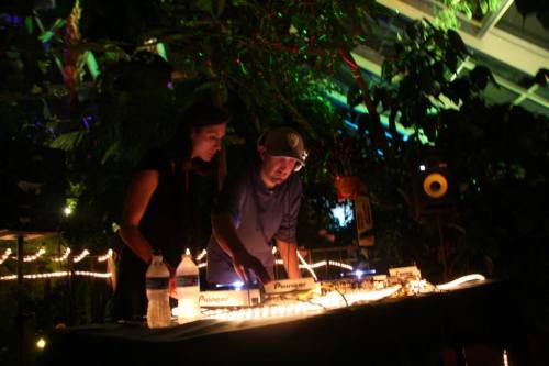 Two DJs perform at a vibrant, illuminated venue surrounded by greenery, focused on their equipment.