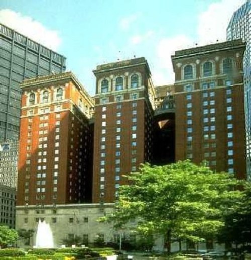 Historic red-brick hotel building with ornate architecture, surrounded by modern skyscrapers and greenery.