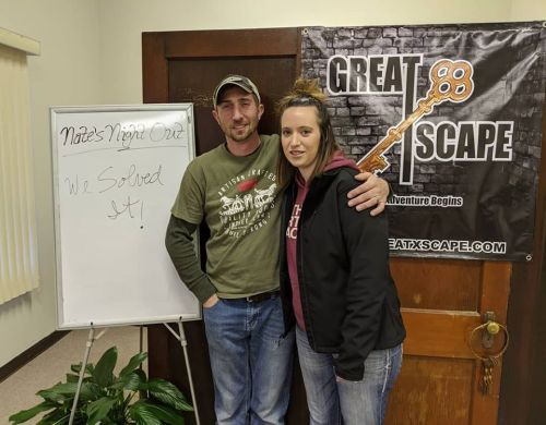 A man and woman stand smiling in front of a sign that reads "Great Escape" with a whiteboard beside them.