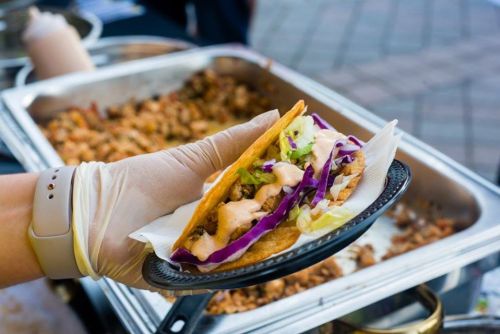 A gloved hand holds a taco with purple cabbage and sauce, with a tray of cooked meat in the background.