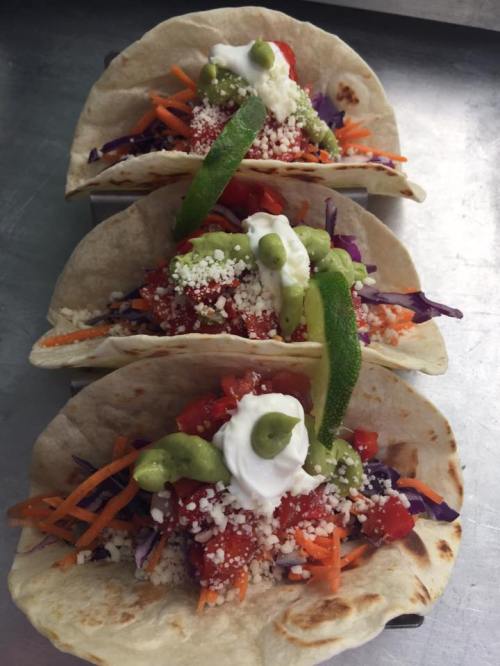 Three tacos filled with colorful vegetables, topped with sour cream, guacamole, and lime wedges on a metal tray.