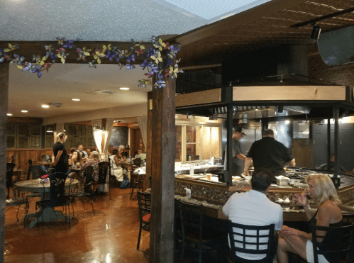 A restaurant interior with diners at tables and chefs cooking in an open kitchen area. Decor includes floral accents.