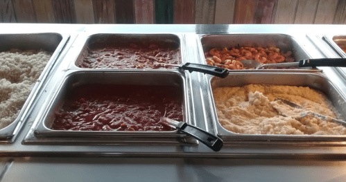 A buffet with four trays: rice, a red sauce, shrimp, and creamy grits, all with serving utensils.