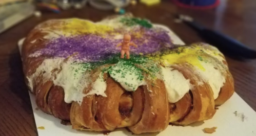 A colorful king cake topped with icing and sprinkles, featuring a small plastic figurine on top.