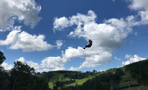 A person zip-lining against a backdrop of blue sky and fluffy clouds, with green hills in the distance.