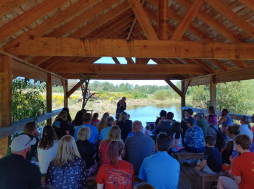 A group of people gathered under a wooden pavilion by a pond, listening to a speaker. Bright greenery surrounds the area.