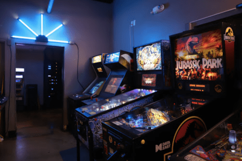 A dimly lit arcade with pinball machines, including a Jurassic Park game, and neon lights in the background.