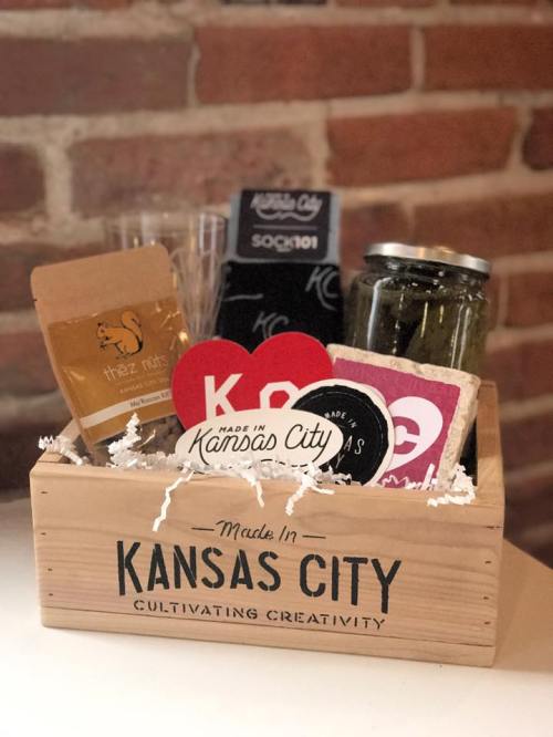 A wooden gift basket filled with local Kansas City products, including snacks, stickers, and a jar, set against a brick wall.