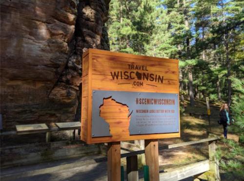 Wooden sign for Travel Wisconsin, featuring the state outline and information about scenic attractions. Surrounded by trees.