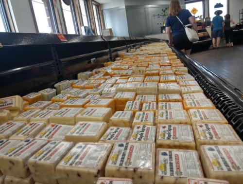 A display of various cheese blocks in a store, with shoppers browsing in the background.