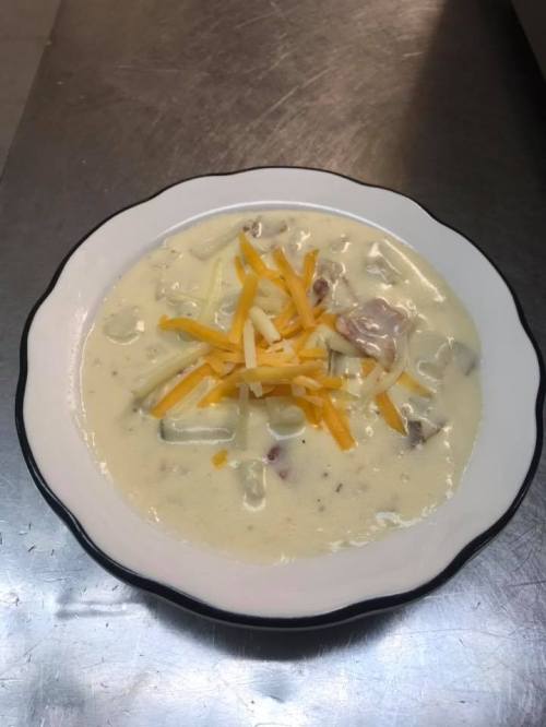 A bowl of creamy soup topped with shredded cheese, featuring chunks of meat and vegetables.
