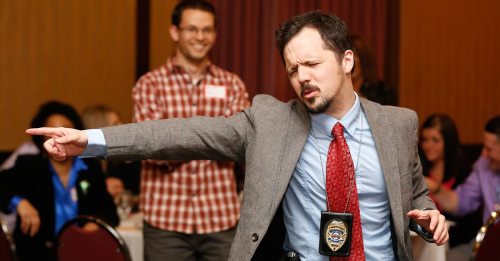 A man in a suit and badge playfully points while dancing, with a smiling man in a plaid shirt in the background.