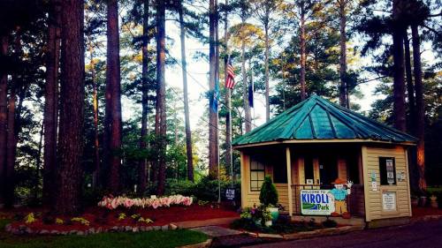 Welcome sign at Kirol Park, surrounded by tall trees and colorful flowers, with an American flag flying nearby.