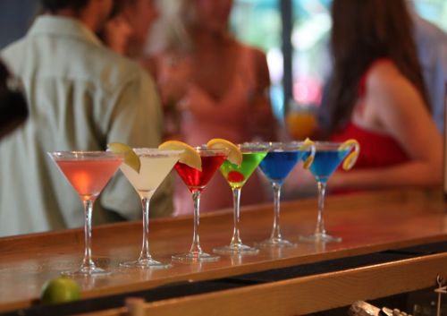 A row of colorful cocktails with lemon slices, set on a bar, with blurred people socializing in the background.
