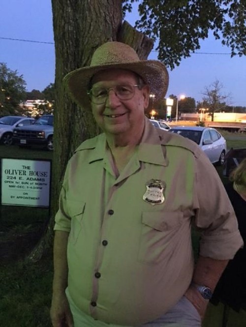 An older man wearing a straw hat and a light-colored shirt stands outdoors near a sign, smiling at the camera.