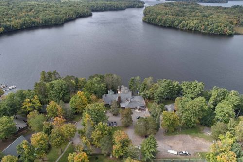 Aerial view of a lake surrounded by trees, with a large house and parking area visible near the water's edge.