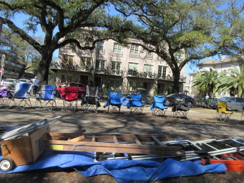 A street scene with parked cars, folding chairs, and a blue tarp under large trees in a sunny area.
