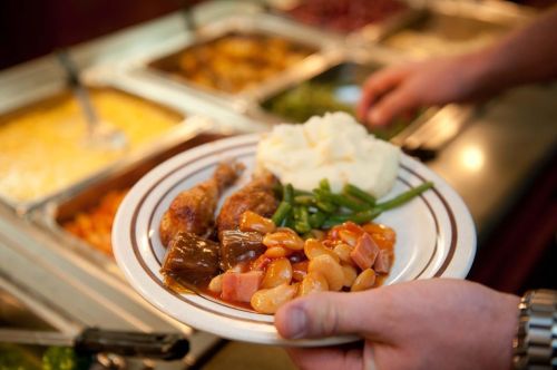 A person holds a plate with chicken, green beans, mashed potatoes, and a mix of beans and ham, with a buffet in the background.