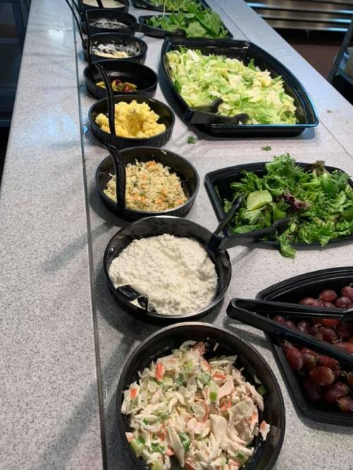 A variety of salads and sides in black bowls on a countertop, including greens, coleslaw, and grapes.