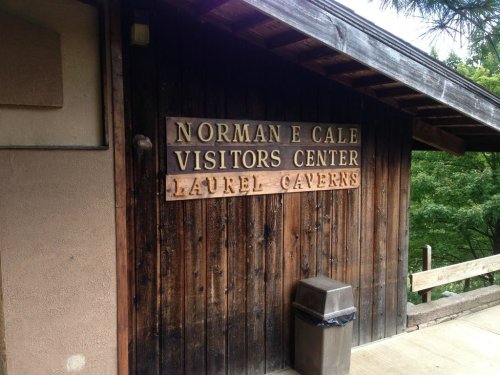 Sign on a wooden building reads "Norman E. Cale Visitors Center, Laurel Caverns" with a trash can nearby.