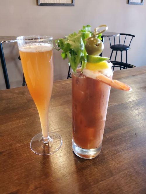 A glass of beer next to a Bloody Mary garnished with shrimp, olives, and lettuce on a wooden table.