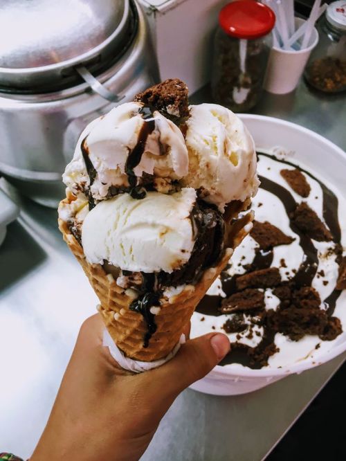 A hand holding a cone with three scoops of ice cream, topped with chocolate sauce, next to a bowl of ice cream.