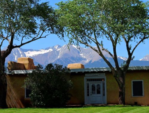 A rustic building framed by trees, with snow-capped mountains in the background under a clear blue sky.