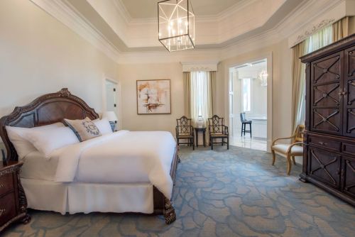 Elegant hotel room featuring a king bed, decorative furniture, and large windows with soft natural light.